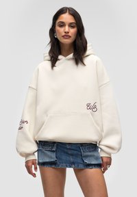 Kremowy, oversize'owy hoodie z kieszenią kangurką. Posiada bordowy haftowany napis na rękawie i przodzie. Można zestawić z dżinsową mini spódnicą.