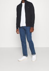 Giacca zip-up scura sopra una maglietta bianca. Jeans blu e sneaker bianche. Presenta un piccolo logo sulla giacca, vestibilità casual e tessuto liscio.