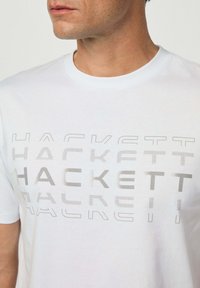 Bílý bavlněný tričko s velkým, texturovaným logem "Hackett" v různých odstínech šedi na přední straně. Klasický design s kulatým výstřihem.