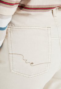 Beige denimbukser med en baklomme med en bølgete brun stingdesign. Tøyet har en subtil tekstur med klare sømdetaljer.