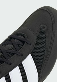 Czarne buty sportowe z siateczkową tkaniną, gładkimi nakładkami, czarnymi sznurowadłami i trzema białymi paskami po bokach. Widoczne szczegóły o teksturowanej powierzchni.