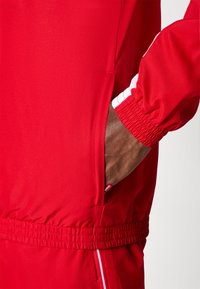 Κόκκινο αθλητικό μπουφάν με υφασματική υφή, ελαστικούς μανικιών και πλαϊνή τσέπη. Λευκές διακριτικές ρίγες κατά μήκος των μανικιών.