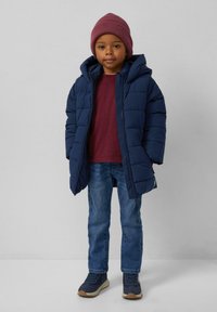 Kind dat een bordeauxrode beanie, een marineblauw pufferjack, een kastanjebruin shirt, een spijkerbroek en marineblauwe sneakers draagt, staat tegen een lichtgrijze achtergrond.