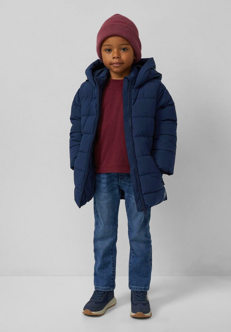 Kind dat een bordeauxrode beanie, een marineblauw pufferjack, een kastanjebruin shirt, een spijkerbroek en marineblauwe sneakers draagt, staat tegen een lichtgrijze achtergrond.