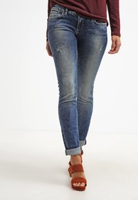 Person som bär uppvikta slitna blå jeans och bronsfärgade sandaler med öppen tå och klack, som går framåt mot en neutral bakgrund.