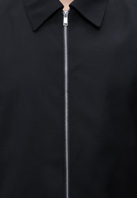 Veste noire avec une fermeture éclair argentée sur toute la longueur et un large col, montrée fermée jusqu’au cou sur une personne.