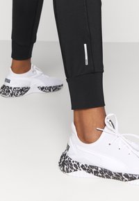 Weiße Sneaker mit einer Leopardenmuster-Sohle, die eine strukturierte Oberfläche und ein Schnürdesign aufweisen; schwarze Leggings mit engem Bündchen.