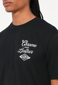 Juodas medvilninis marškinėlis su tekstūruota balta grafika, kurioje užrašyta "Chrome and Leather" ir "Speed Replay Shop" deimanto forma. Trumpomis rankovėmis.