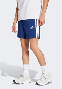 Blauwe sportshorts met witte zijkanten, gecombineerd met witte sokken en lichtgekleurde sneakers. De shorts hebben het Adidas-logo.