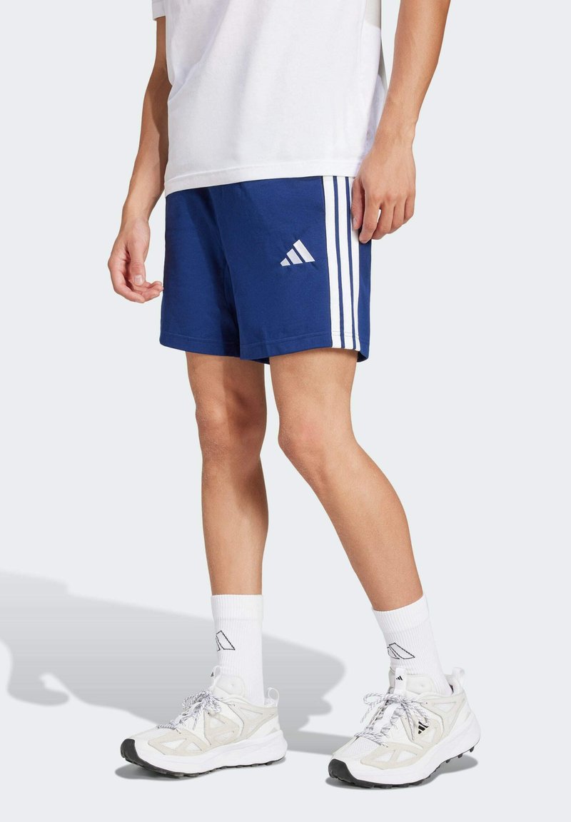 Blauwe sportshorts met witte zijkanten, gecombineerd met witte sokken en lichtgekleurde sneakers. De shorts hebben het Adidas-logo.