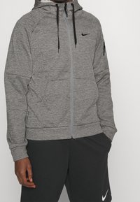Hombre vestido con una sudadera gris con cremallera y cordones negros, y pantalones cortos negros de Nike, de pie con una mano en el bolsillo frente a un fondo blanco.