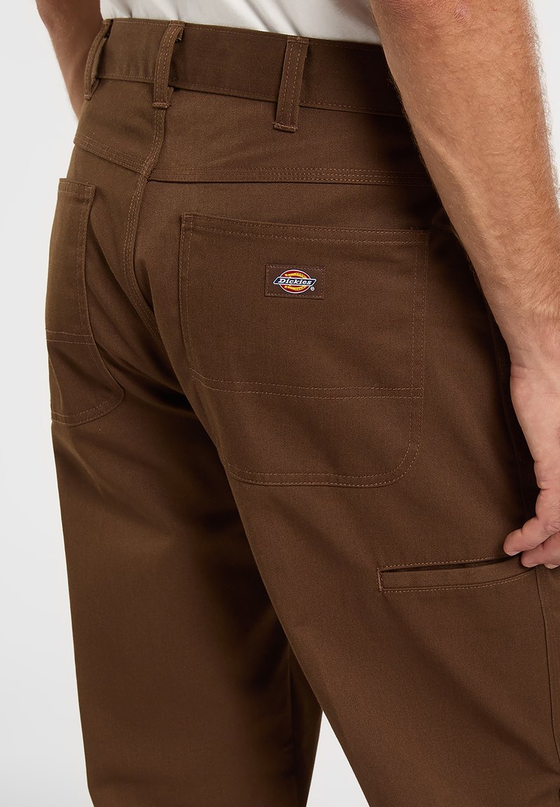 Brown work pants with a solid texture, featuring back pockets and a visible brand patch. The design includes belt loops and sturdy seams.