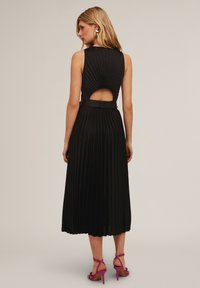 Schwarzes Faltenkleid mit einem ausgeschnittenen Rücken, taillierter Brust und einem Rock, der bis zur Mitte der Wade reicht. Kombiniert mit leuchtend pinken, strappy Heels.
