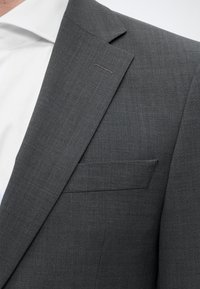 Gros plan sur le revers et la poche poitrine d'une veste de costume grise portée sur une chemise blanche.