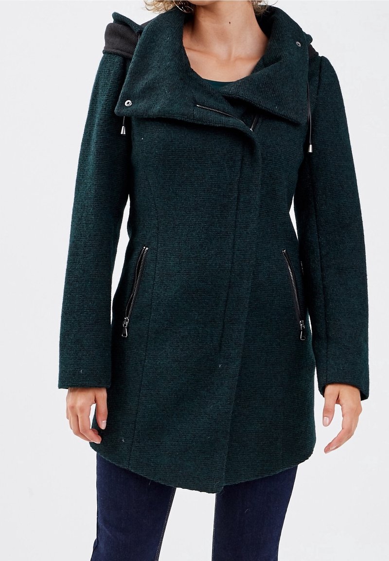Femme portant un manteau en laine vert foncé avec un col asymétrique et des poches latérales zippées, associé à un pantalon bleu foncé.