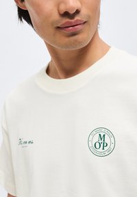 Άνδρας που φοράει λευκό μπλουζάκι με πράσινο κείμενο «It's on us.» και κυκλικό λογότυπο «FC Marc O'Polo Stockholm 1967» στο στήθος.