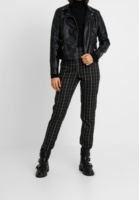 Sort lædermotorcykeljakke, tætsiddende sort rullekrave og sort-hvide ternede, tilspidsede bukser med knæhøje sorte støvler med spænder.