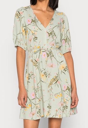 Kobieta ubrana w jasnozieloną sukienkę w kwiaty z krótkimi bufkowatymi rękawami, zapinaną na guziki z dekoltem w kształcie litery V oraz przewiązanym paskiem w talii.