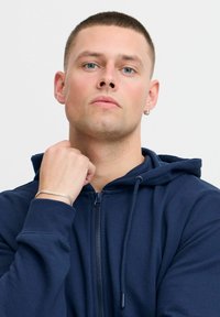 Marineblauwe zip-up hoodie met een trekkoord kap, zachte textuur en ritssluiting aan de voorkant. Gecast tegen een neutrale achtergrond, gedragen met een zilveren kettingarmband.