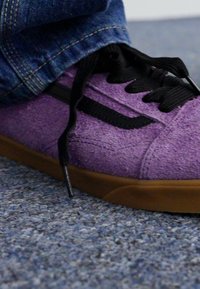 Lila velúr sneaker fekete fűzőkkel, texturált felső résszel és barna gumi talppal. Farmernadrág részben látszik a cipő felett.