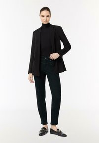 Zwarte blazer over een zwarte coltrui, gecombineerd met donkergroene broek en zwarte instapschoenen met gouden hardware accenten. Soepele, getailleerde pasvorm.