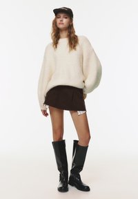 Mladá žena v krémovom oversized svetri, tmavej mini sukni, čiernych vysokých čižmách a čiernom klobúku, postávajúca pred obyčajným bielym pozadím.