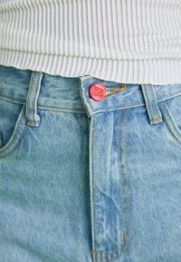 Jean en denim bleu clair avec un bouton rouge arborant le logo "be", design standard à cinq poches et coutures jaunes contrastantes.