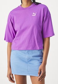 Fialové zkrácené tričko s krátkým rukávem, na levé části hrudi s bílým logem Puma. Kombinováno s světle modrou džínovou sukní.
