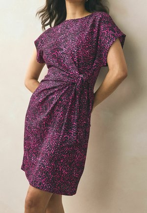 Mujer con un vestido corto de manga corta hasta la rodilla con estampado de leopardo en púrpura y negro, de pie contra una pared beige.