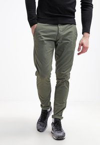 Úzké olivově zelené chinos s hladkou texturou, které mají klasický design s pěti kapsami a ohrnutými manžetami. Černé sportovní boty doplňují celý outfit.