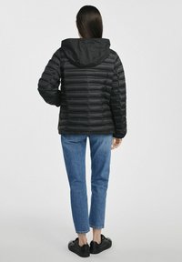 Czarna kurtka puchowa z kapturem, z poziomym pikowaniem. W zestawieniu z niebieskimi dżinsami i czarnymi butami. Widok z tyłu.