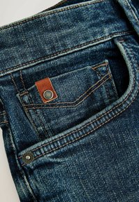 Tumed sinised teksad tekstuuriga pinnaga, millel on pruun nahkplaat ja metallnööbid taskul.