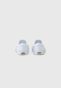 Valged kanvasest slip-on kingad madala profiiliga, kummist tallaga ja musta logoga kanna peal. Sujuv tekstuur ja lihtne disain.