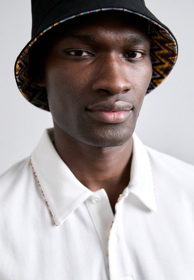 Λευκή μπλούζα πόλο με υφασμάτινο γιακά που διαθέτει πολύχρωμες λεπτομέρειες, σε συνδυασμό με ένα μαύρο καπέλο μπucket με επένδυση σε ζιγκ ζαγκ μοτίβο.