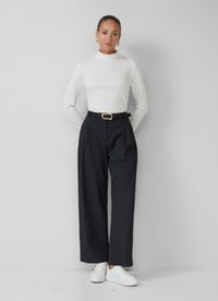Femme debout contre un mur uni, portant un haut blanc à col montant, un pantalon large à rayures noires avec une ceinture et des baskets blanches.