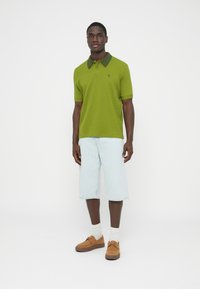 Zöld galléros póló sötét gallérral, világoskék farmer rövidnadrág, barna lábbeli és fehér zokni. Egyszerű, texturált anyag és laza szabás.