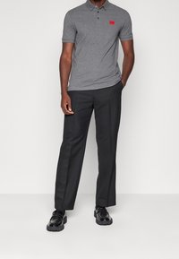 Camisa polo gris con un logo rojo, combinada con pantalones negros y zapatos negros. El tejido de la polo parece suave y transpirable.
