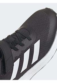 Gros plan sur une chaussure de sport noire avec des rayures blanches en diagonale, des lacets noirs et une semelle texturée blanche.