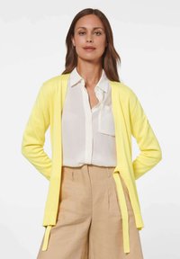 Geel gebreide cardigan met een lichte textuur, open voorkant en strikaccenten, gecombineerd met een wit shirt en beige broek.