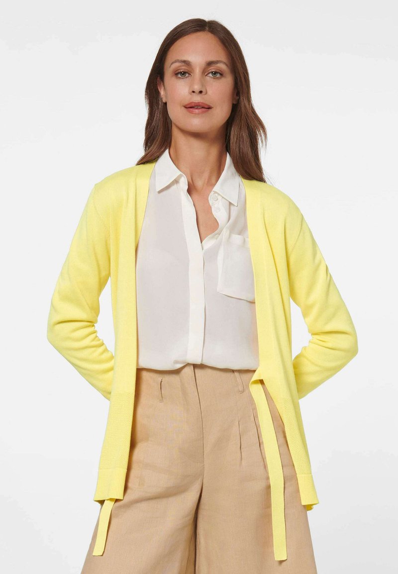 Geel gebreide cardigan met een lichte textuur, open voorkant en strikaccenten, gecombineerd met een wit shirt en beige broek.