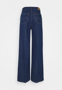 Jean en denim bleu foncé à jambes larges vu de dos avec deux poches plaquées et un patch de marque en cuir marron à la taille.