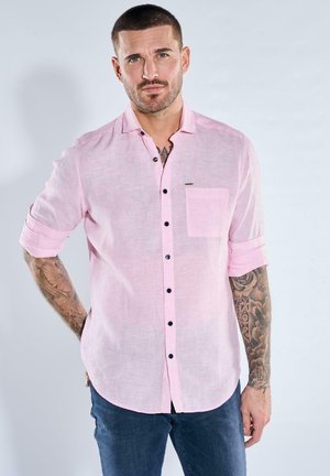 Mann mit Tätowierungen, der ein hellrosa Hemd mit hochgekrempelten Ärmeln und dunkelblaue Jeans trägt, steht mit der Hand am Rücken.