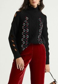 Pull noir en maille avec épaules découpées, orné de motifs circulaires colorés et d'un motif en losange texturé. Associé à un pantalon en velours rouge.