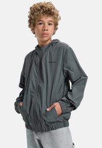 Grå lättviktsvindjacka med huva, framdragkedja, elastiska ärmmuddar och sidofickor. Slät textur med minimal logotyp.