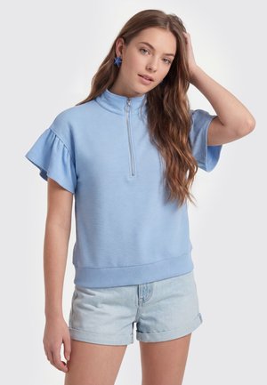 Jeune femme aux longs cheveux bruns portant un haut bleu clair à manches courtes avec une fermeture éclair et un short en jean, touchant ses cheveux d'une main.