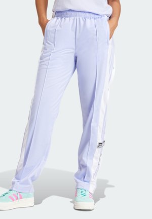 Person trägt hellviolette Adidas-Trainingshose mit weißen Seitenstreifen und türkisfarbene Sneaker mit pinken Akzenten, Hände in den Taschen.