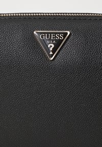 Portafoglio in materiale nero testurizzato con logo triangolare di Guess U.S.A. decorato da un punto interrogativo, accentuato da hardware e zip in tonalità dorata.