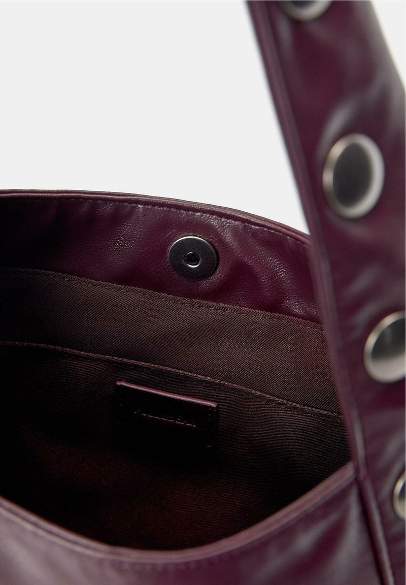 Bolso de cuero morado oscuro abierto con forro de tela oscura y cierre magnético plateado en el borde interior del bolso.