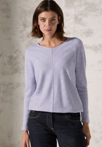 Helllila gestrickter Pullover mit einem Zickzackmuster, Rundhalsausschnitt und langen Ärmeln, kombiniert mit dunklen Jeans. Strukturierte Stoffdetails sind sichtbar.