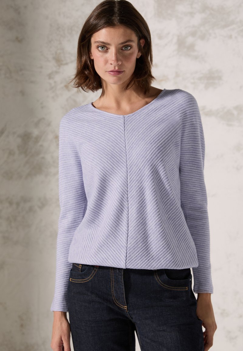 Helllila gestrickter Pullover mit einem Zickzackmuster, Rundhalsausschnitt und langen Ärmeln, kombiniert mit dunklen Jeans. Strukturierte Stoffdetails sind sichtbar.